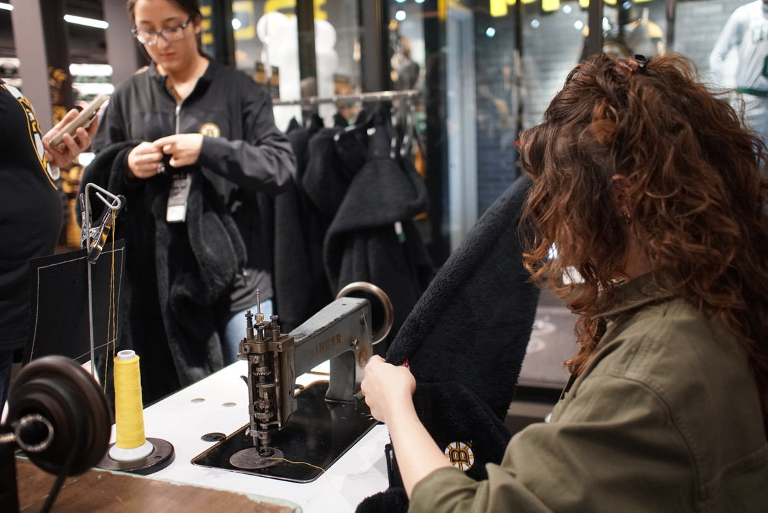 A woman with curly brown hair sitting behind a vintage singer chainstitch embroidery machine. She is holding up a fleece garment with the Boston Bruins Logo on it. The location of the picture is at the TD garden pro shop in Boston.