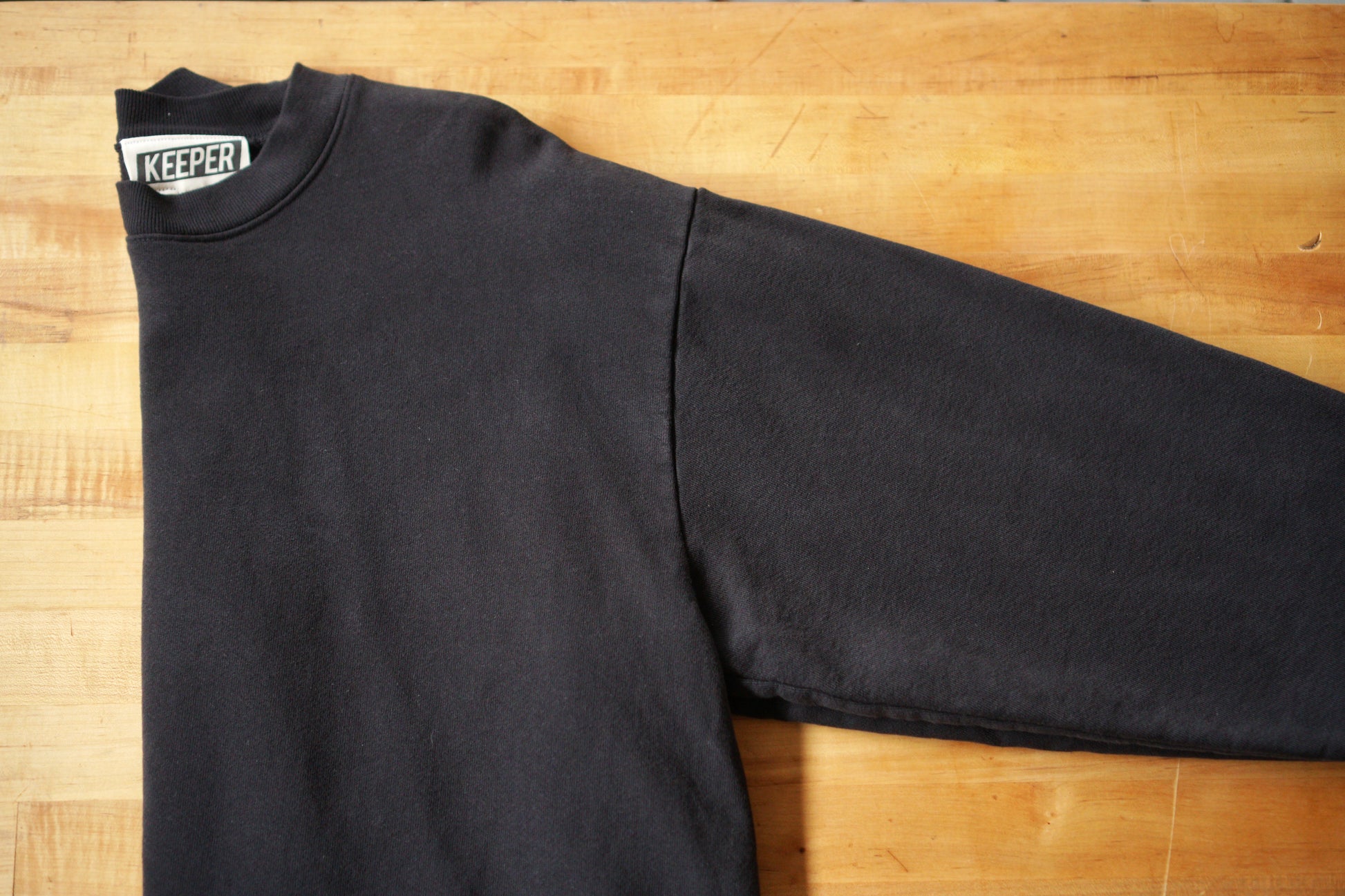 A black crewneck sweatshirt with a visible tag that says KEEPER. This sweatshirt has a chainstitch embroidered embellishment in a cursive font on the left chest area.