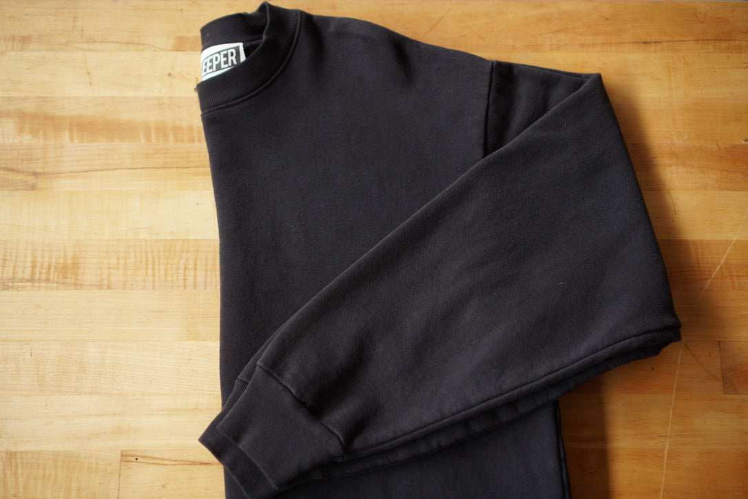 A folded black crewneck sweatshirt with a visible tag that says KEEPER. This sweatshirt has a chainstitch embroidered embellishment in a cursive font on the left chest area.