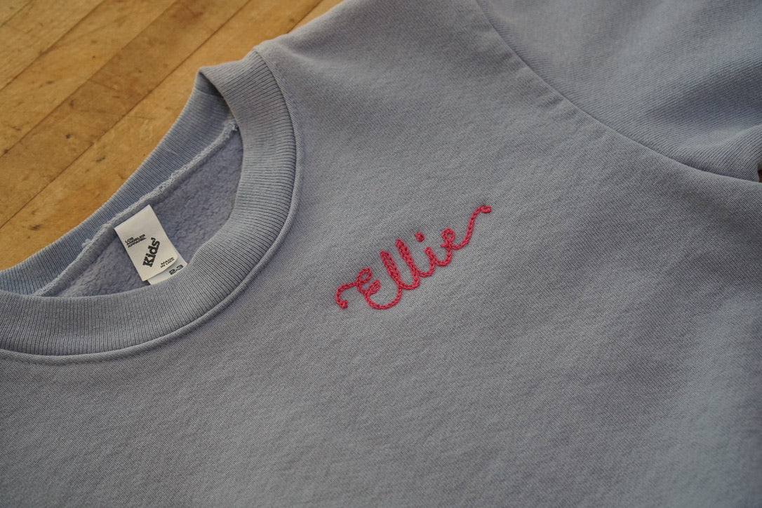 A light blue sweatshirt with the name "Ellie" embroidered in a pink thread. The name is in a cursive / script font and was done using a vintage chainstitch machine.