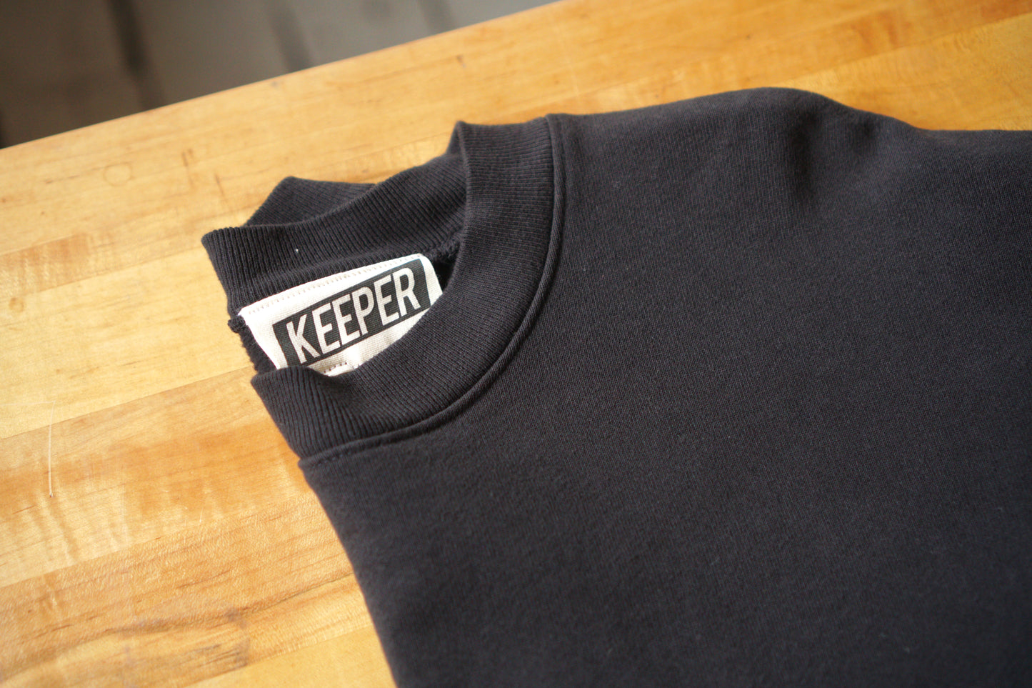 A close up of the neckline of a black crewneck sweatshirt with a visible tag that says KEEPER. This sweatshirt has a custom chainstitch embroidered embellishment in a cursive font on the left chest area.