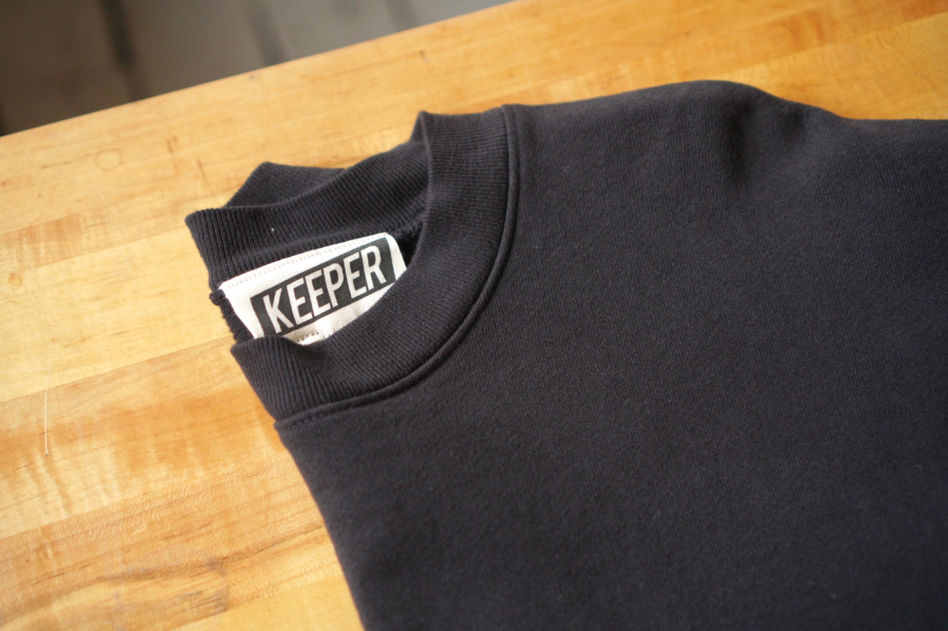 A close up of the neckline of a black crewneck sweatshirt with a visible tag that says KEEPER. This sweatshirt has a custom chainstitch embroidered embellishment in a cursive font on the left chest area.
